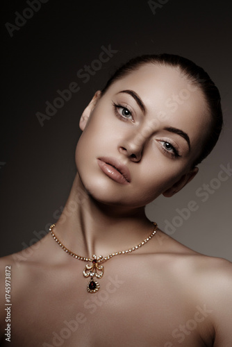 Elegant close-up portrait of a woman with nude makeup and a gold necklace with gemstone pendant. Fashion and beauty concept with soft studio lighting and neutral background.
