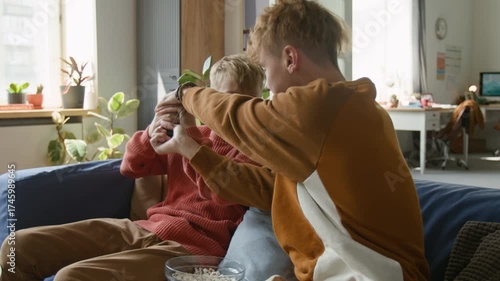 Medium shot of young teenage boy fighting with brother for TV remote while sitting on couch together and eating popcorn