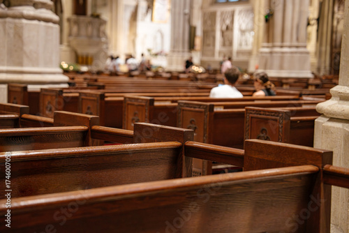 Wooden church pews fill the foreground, leading to a softly lit cathedral interior. A few people are seated, creating a quiet and reverent atmosphere in the sacred space.