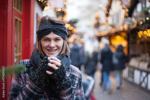 Beautiful woman holding a cup of mulled wine, while standing on traditional Christmas market in Germany. Portrait of cheerful model in winter outfit enjoying  cup of Christmas holiday spirit.