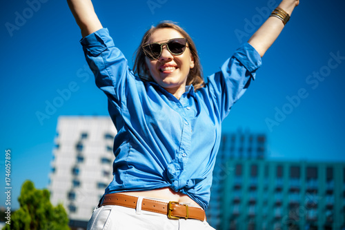 Young woman with blonde hair smiles brightly while celebrating outdoors in a blue shirt, surrounded by modern buildings under a clear sky.