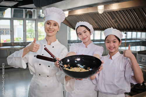Teenage trainee girls in white aprons are proud to show fried rice in pan, cooking class work with master chef in gourmet restaurant kitchen, delicious cuisine with ingredients, and culinary meals.