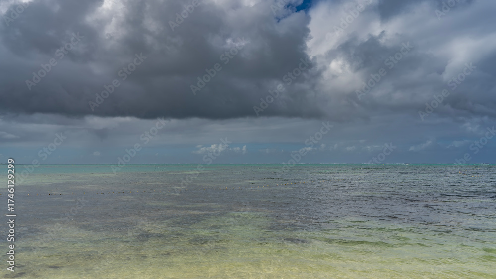 Obraz premium Beautiful seascape under a cloudy sky. Turquoise ocean, ripples on the surface of the water, foam waves on the horizon. There are dark clouds in the blue sky. Mauritius.