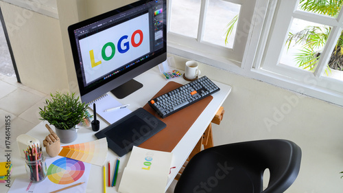Top view of graphic designer desk with computer screen showing colorful logo design, drawing tablet, and color palette