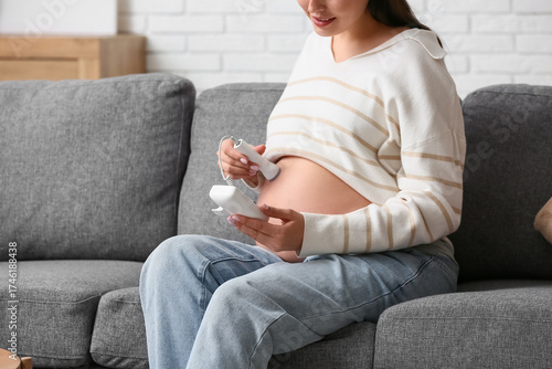 Fotografie Young pregnant woman with fetal doppler listening to baby's heartbeat on sofa at