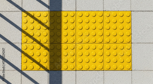 Yellow tactile paving blocks arranged on a concrete surface, casting shadows from nearby structures, enhancing urban accessibility and safety for pedestrians navigating the area