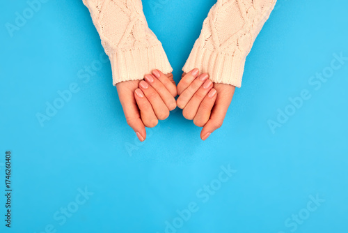 Woman's hands wearing a cozy cream knitted sweater displaying perfectly manicured nude nails on a vibrant blue background, creating a comforting winter beauty concept