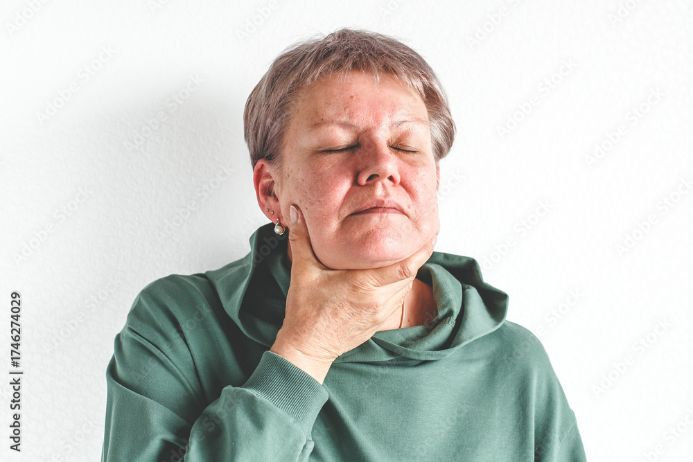 Obraz premium Middle-aged woman in green sweatshirt touching her throat with painful expression, symbolizing sore throat, thyroid gland disorder, inflammation or respiratory illness and health awareness
