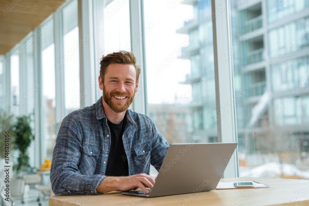 Fototapeta premium A happy man sitting at a table, using a laptop in a bright, modern home office. the man he is smiling while working online or looking for inspiration.