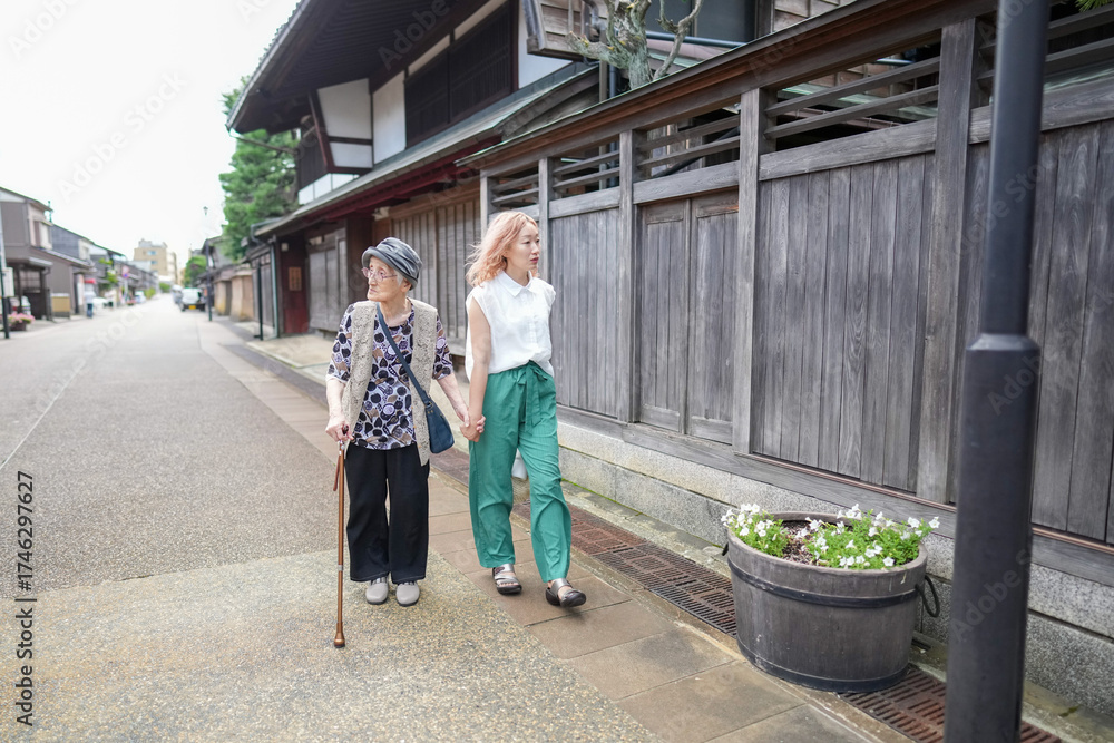Fototapeta premium Two older women are walking down a street, one of them holding a cane. They are both wearing green pants and white shirts. The street is lined with wooden buildings, giving the scene a quaint