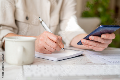 Woman managing personal finance, writing down expenses in a notebook while holding phone representing online banking or financial applications, focusing on home economy tasks
