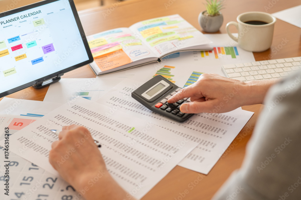 Fototapeta premium Person calculating finances at a desk with calculator, pen, financial documents, charts and tablet calendar, managing budgeting, expenses, taxes and planning for business or personal goals