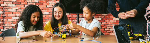 Three girls collaborate in classroom lab, assembling wheeled robot with wires. Guided by teacher, students engage in programming lesson, showing teamwork, creativity, and hands-on learning.