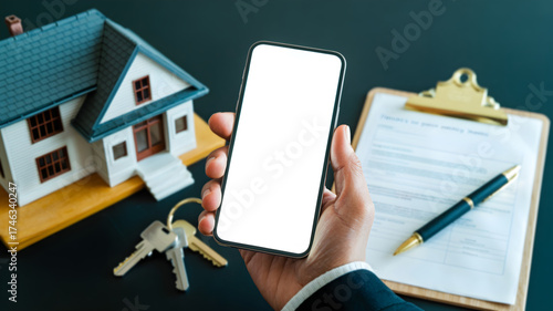 Hand holding a smartphone with a blank white screen, next to a model house and contract documents