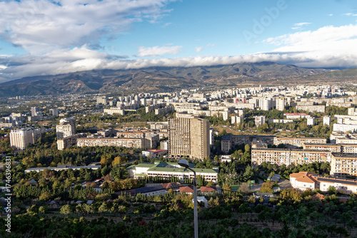 The scenery of Tbilisi city , Georgia, with a lot of buildings and trees. The sky is cloudy and the mountains are in the backgorund.