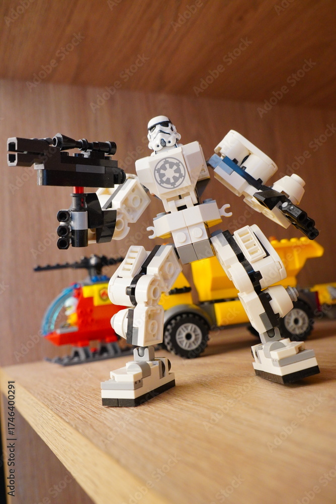 Naklejka premium Custom-built LEGO Star Wars Stormtrooper mech robot figure holding a blaster weapon, displayed on a wooden shelf with other toys in the background