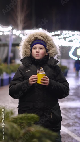 Vertical portrait of boy dancing with sweet pastries at winter Christmas market in evening. Child in warm jacket stays warm on cold, frosty evening on street decorated with fairy lights.