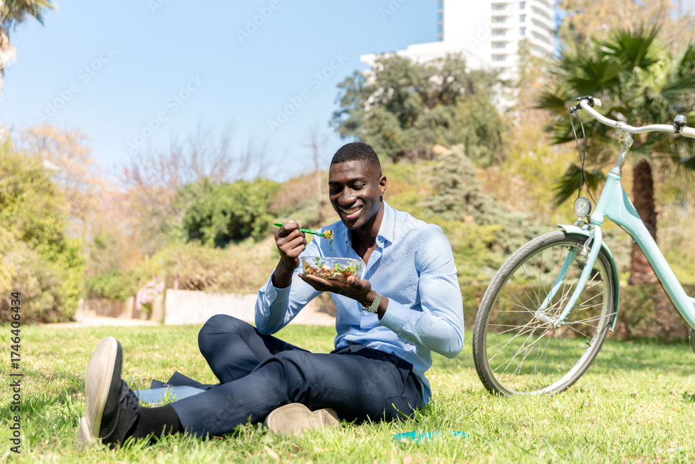 Obraz premium African american Businessman eating salad in park next to his bicycle during lunch break