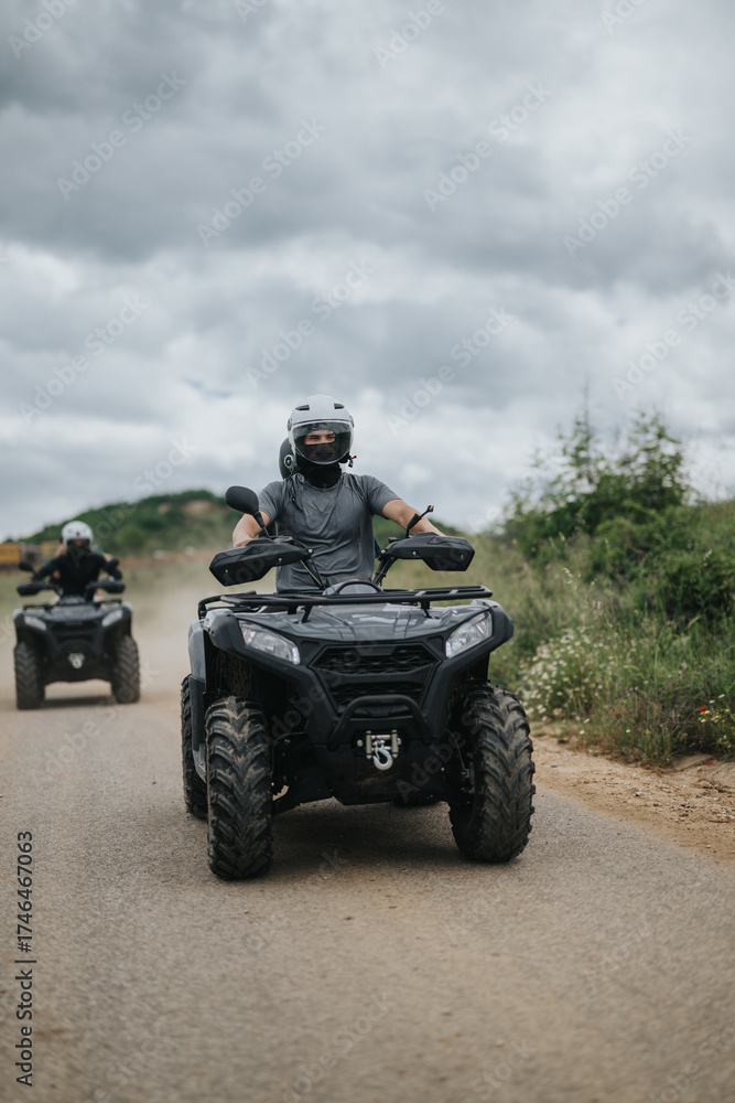 Fototapeta premium Two individuals, equipped with helmets, riding ATVs along a road surrounded by a green landscape, under an overcast sky, embarking on an outdoor adventure.