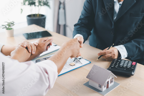 Close up of real estate agent and client shaking hands with house model, contract paperwork and keys on desk, concept of successful property deal, mortgage loan agreement and business partnership.