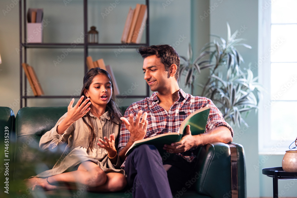 Fototapeta premium Indian father reading book to daughter, sharing quality bonding time on sofa