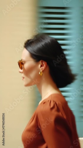 A young Caucasian woman with short black hair walks past a blue shutter. She wears sunglasses and a brown blouse, exuding a sense of style and confidence.