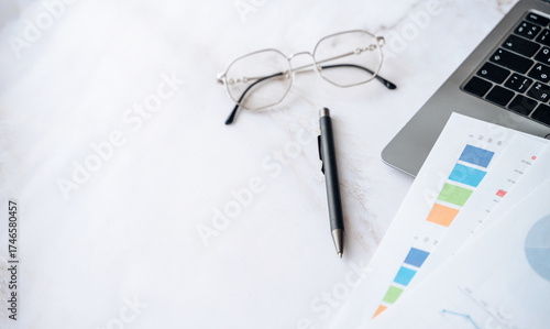Desk setup with glasses, pen, laptop, and financial papers showing charts and graphs for business analysis