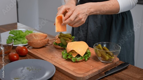 Unrecognizable woman assembling a juicy cheeseburger with patty, sauce, pickles, and lettuce in her kitchen.