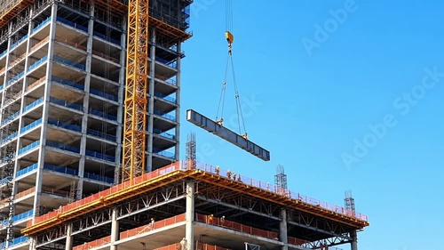 A large crane lifts a long steel beam onto a multi-story building under construction against a clear blue sky.