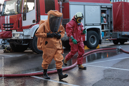 Two emergency responders in full chemical or biohazard suits and breathing apparatus walking in front of a red fire truck at a hazardous material incident scene.