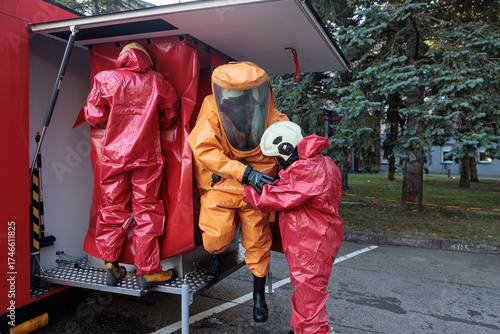 Emergency team members in red chemical suits helping a third responder in an orange hazmat suit enter a mobile decontamination unit or vehicle.
