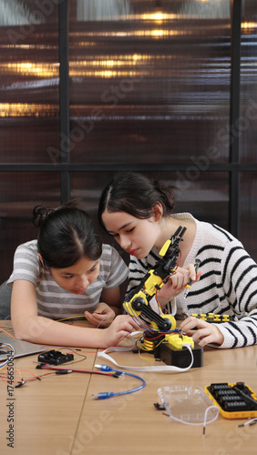 Asian and Caucasian girls collaborate on robotics project, assembling robot parts and sharing ideas with smiles in classroom.
