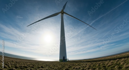 Tall wind turbine stands on grassy coast, bright sun, and streaky sky
