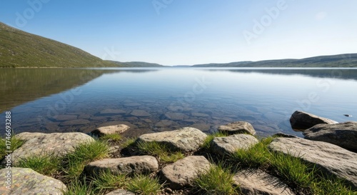Tranquil lake scene with clear water, rocks, and distant mountains under blue sky