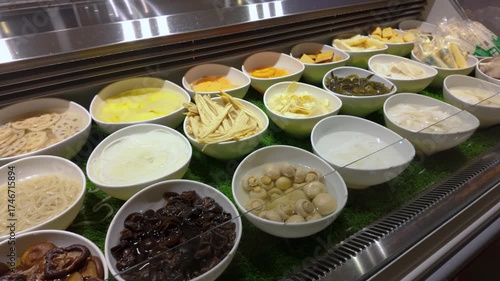 A buffet style food counter displaying various ingredients arranged in white bowls. The selection includes mushrooms, lotus root slices, noodles, tofu, and etc typically found in steamboat restaurant.