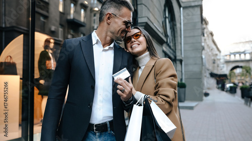 Stylish couple walking in city for shopping, smiling and hugging. Man in dark suit, woman in camel coat and sunglasses holding bags and credit card. Love and luxury lifestyle. Black friday sales