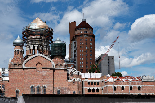 Historic brick church under restoration with scaffolding and copper domes, surrounded by modern buildings under a blue sky. Urban architecture contrast.
