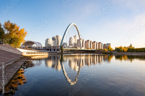 Karaotkel Bridge in Astana Kazakhstan with modern buildings and autumn trees reflected in calm Ishim River
