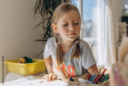 Little girl with pigtails sitting at a table drawing with colored pencils in a bright room. Child focused on creative activity in a cozy home environment
