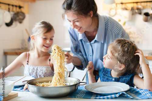 Happy mother and children eating pasta together at home kitchen, enjoying family lunch, healthy comfort food for kids. Homemade Italian cuisine. Candid, authentic lifestyle of solo motherhood