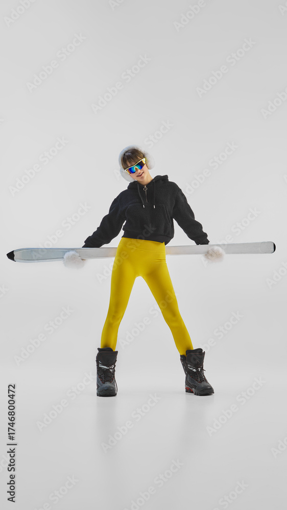 Obraz premium Young woman in bright winter outfit holding skis horizontally with attitude. Concept of influencer winter fashion, confidence, and bold expression for social media, lifestyle, and athleisure branding.