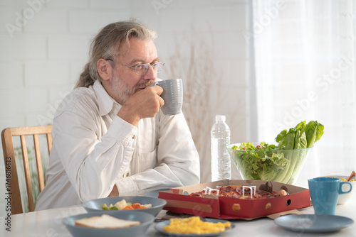Senior elderly older grandfather stay at home with painful face sitting alone on eating table in house. Depressed elderly retired man upset feeling unhappy. Lonely and missing his family at home	
