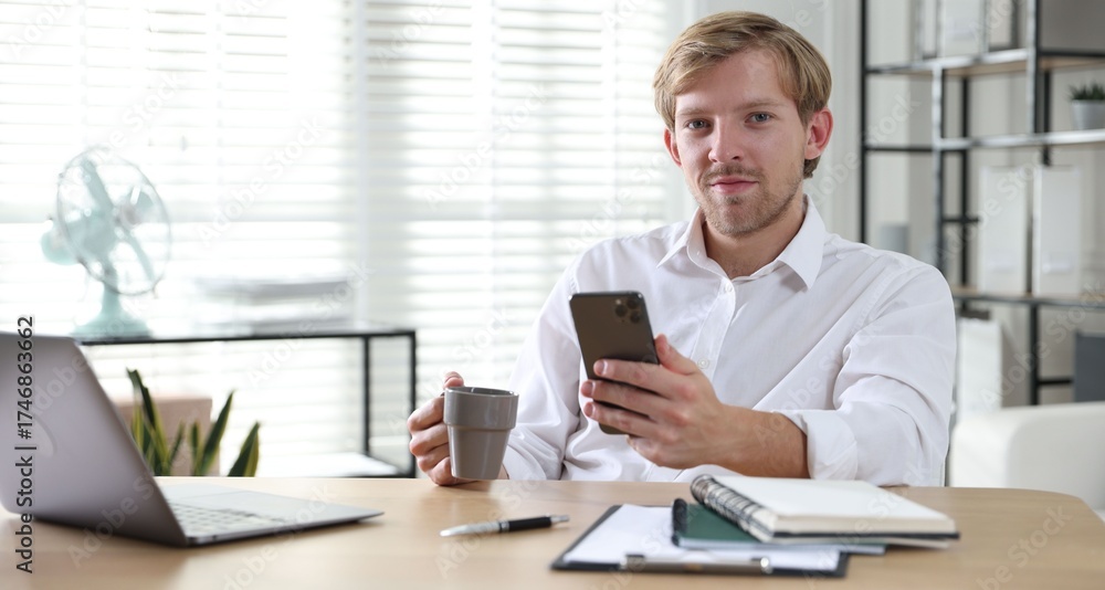 Fototapeta premium Man with cup of drink and smartphone at wooden table indoors