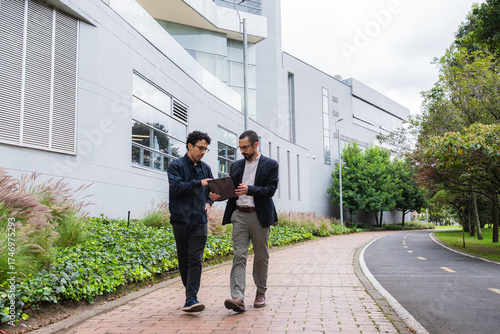 Two men walking and collaborating while viewing a laptop screen in an outdoor setting