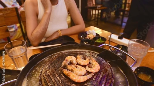 Woman cooking prawns on Thai Moo Krata BBQ hotpot grill in Bangkok restaurant, combining soup and barbecue cooking style with seafood, chopsticks, and dipping sauces at night