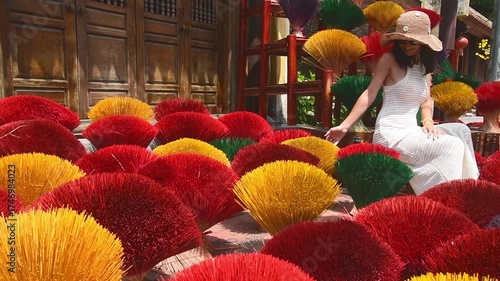 Woman in white dress and straw hat sitting among colorful incense stick bundles at traditional craft village inVietnam, cultural heritage and tourism concept