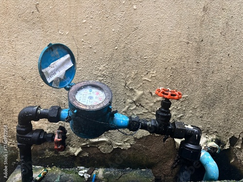 Close-up of an old, worn blue water meter and connecting pipes with rusty valves mounted against a rough, weathered stucco wall, highlighting aging utility infrastructure.