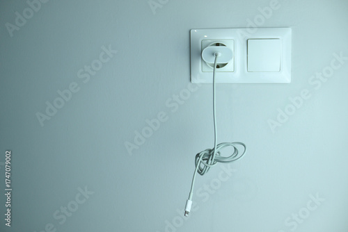 Closeup of white wall socket with charging cable hanging down. Concept of household power, electricity, minimal design, and everyday technology.