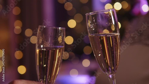 Two champagne glasses clink together in evening in decorated room with garland and warm lighting. Close up of festive glasses in cozy, festive atmosphere.