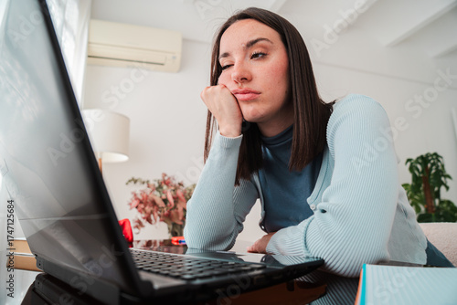 A young woman looking frustrated while working on her laptop at home, showing signs of sadness and stress on her face as she attempts to concentrate on her studies and tackle her academic tasks
